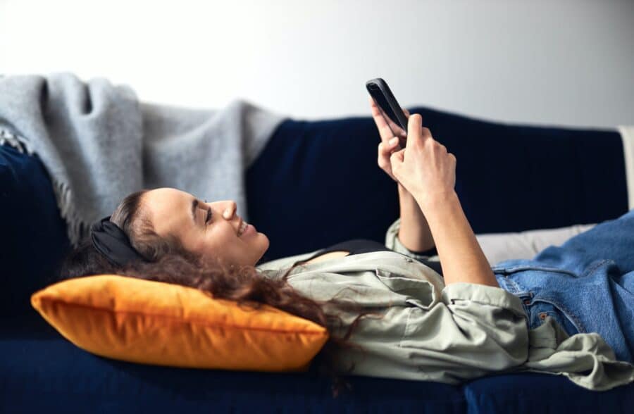 Young Smiling Woman Relaxing At Home Lying On Sofa Checking Social Media On Mobile Phone